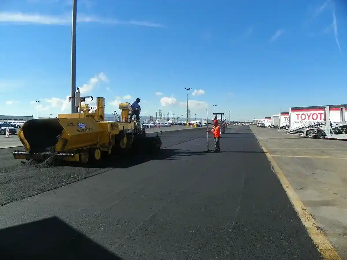 Asphalt paving machine laying fresh pavement on large commercial parking lot with workers on site